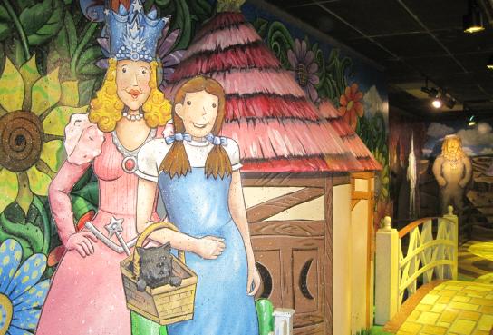 Glinda, Dorothy and Toto in front of munchkinland image of house and oversized flowers