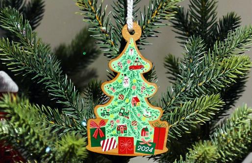 Ornament cut in shape of a Christmas Tree, painted green with ornaments & packages, hanging on evergreen branch