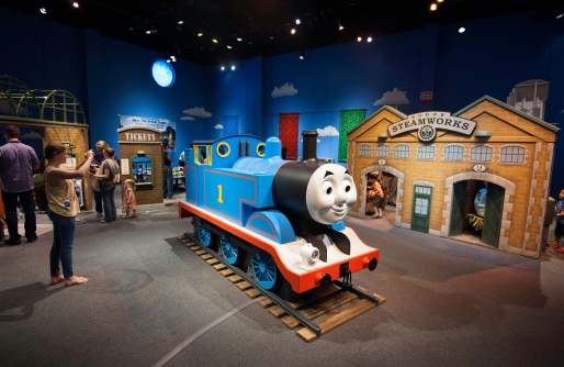 Blue Thomas Engine in front of Village Flat Structures, woman taking picture