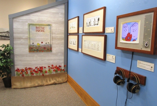 View of Battle Prose content on Barn wall with Poppies and frames cartoon artwork on blue wall next to TV screen with headphones featuring an image of Charlie Brown and Linus